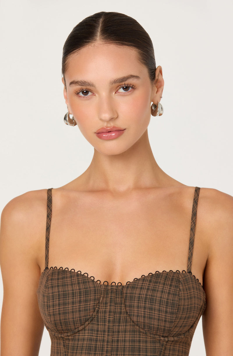 A young woman with fair skin and dark brown hair styled in a sleek, middle-parted bun. She is wearing large, silver hoop earrings. Her outfit is a brown, plaid-patterned bustier top with thin, matching spaghetti straps. The top features a scalloped trim along the sweetheart neckline.