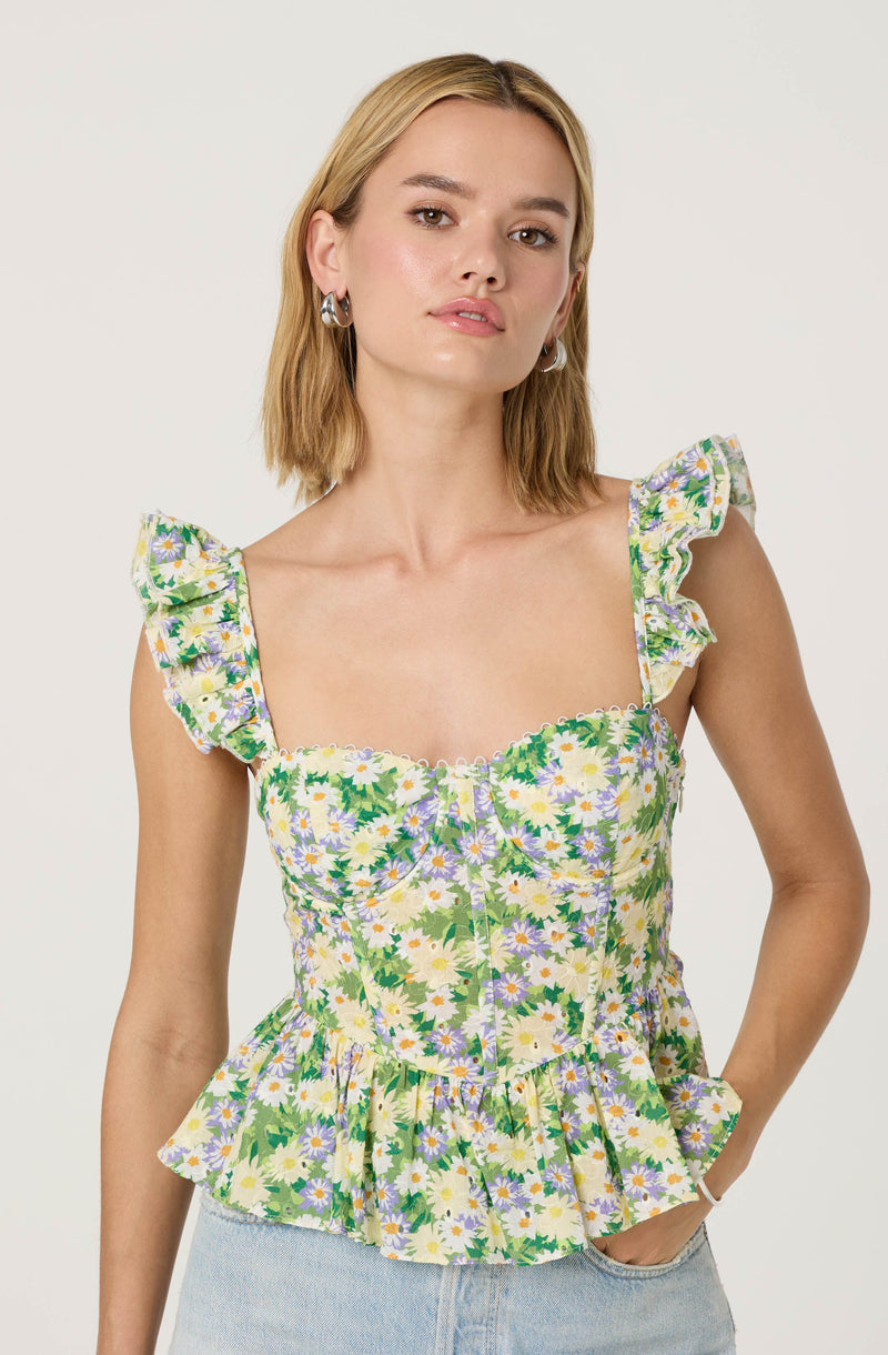 A woman is wearing a sleeveless, fitted top with ruffled shoulder straps. The top features a colorful floral pattern with daisies and green leaves on a light background. The hem of the top has a ruffled peplum style.