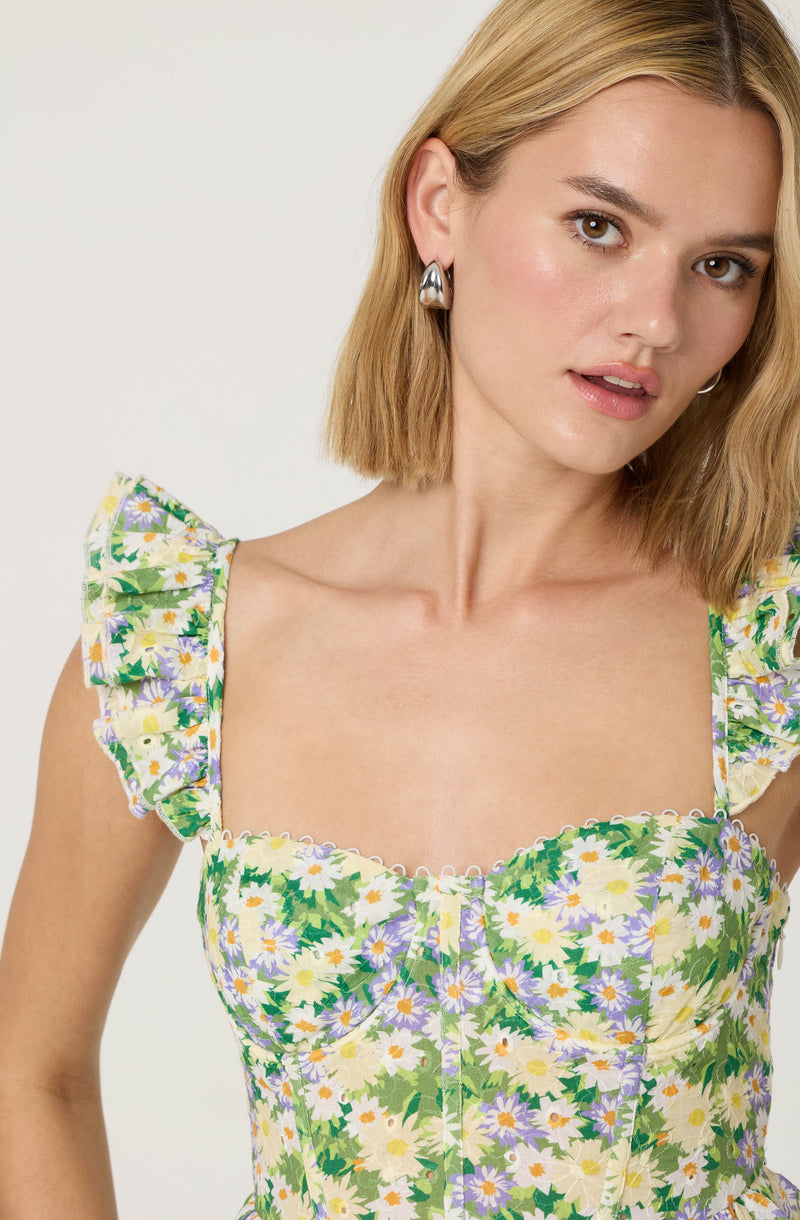 A woman is wearing a sleeveless dress with a floral pattern. The dress features colorful daisies and purple flowers with green foliage, set against a light background. The shoulder straps are ruffled, adding a decorative touch to the dress.