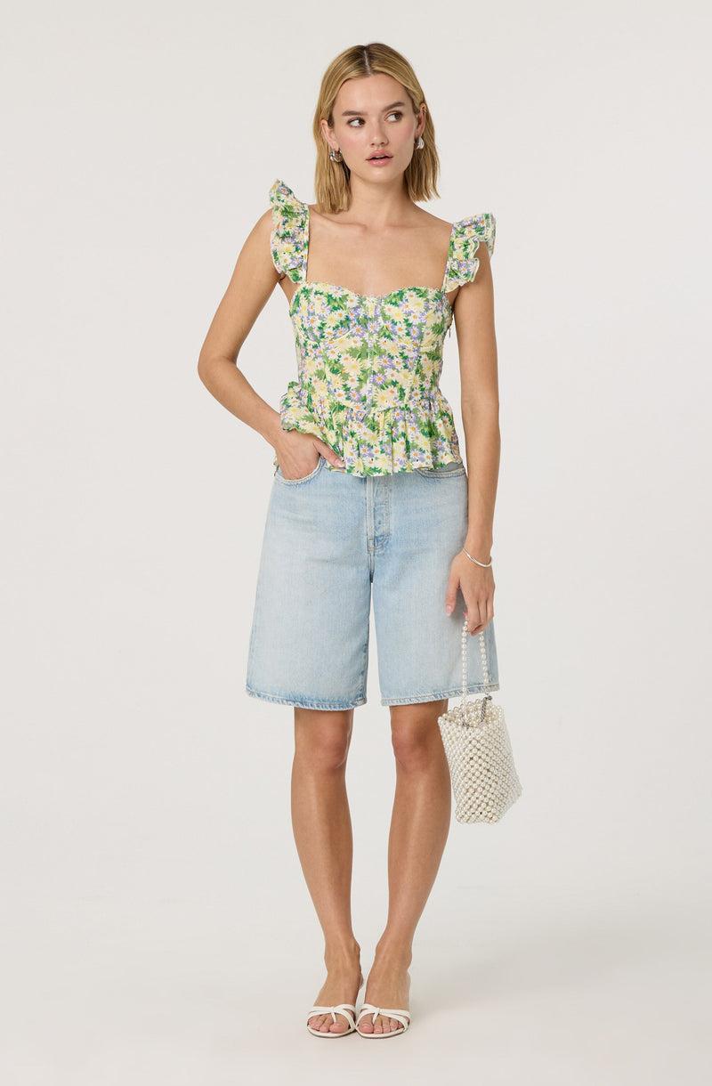 A woman is wearing a sleeveless, floral-patterned top with ruffled shoulder straps and a peplum waist. She is paired with light denim knee-length shorts. She has on white sandals with thin straps and is holding a small, white, woven handbag with a pearl handle.