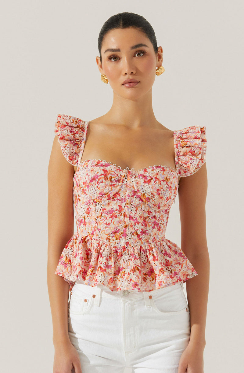 The woman is wearing a sleeveless, fitted top with ruffled shoulder straps. The top has a floral pattern in shades of pink, orange, and white, with a ruched bodice and a peplum-style ruffle at the hem. She is also wearing a pair of white pants.