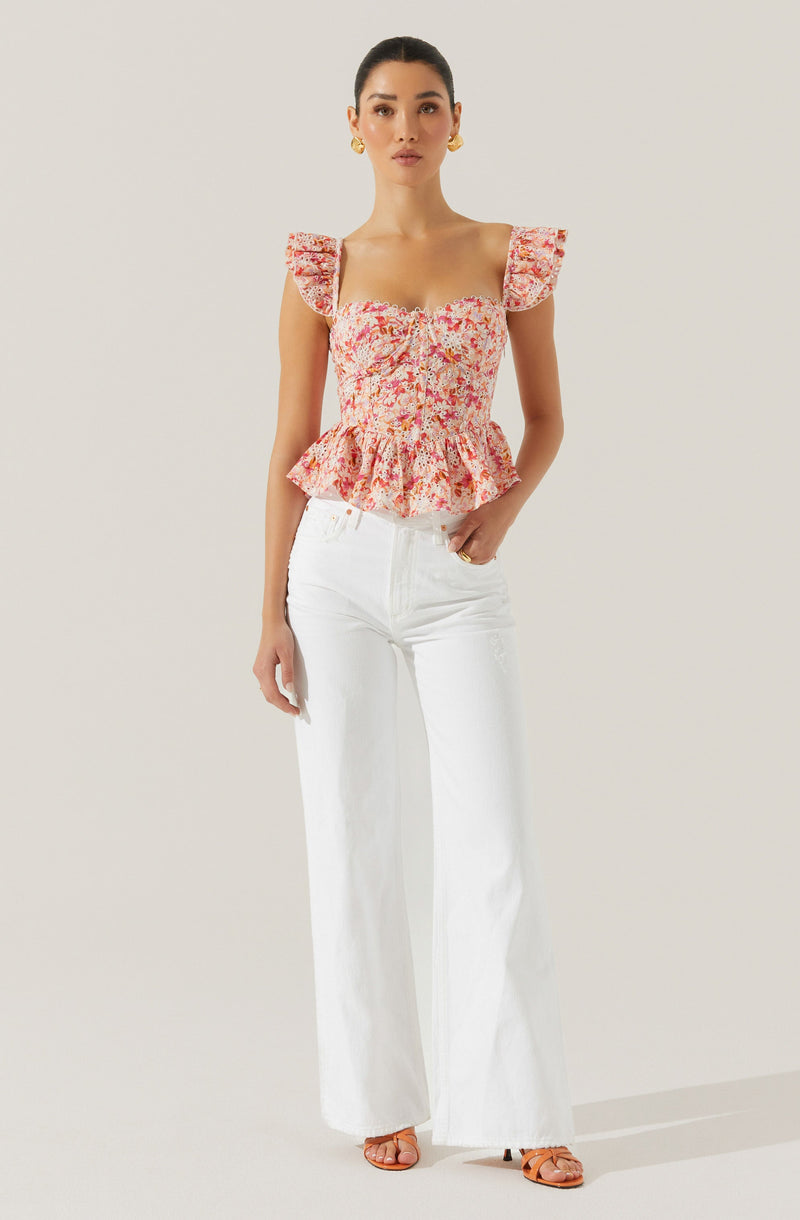 A woman is wearing a pink floral top with ruffled shoulder straps and a gathered, peplum-style hem. She pairs it with high-waisted, white wide-leg jeans and open-toed, strappy sandals. She has gold hoop earrings and her dark hair is styled in a sleek, pulled-back look.