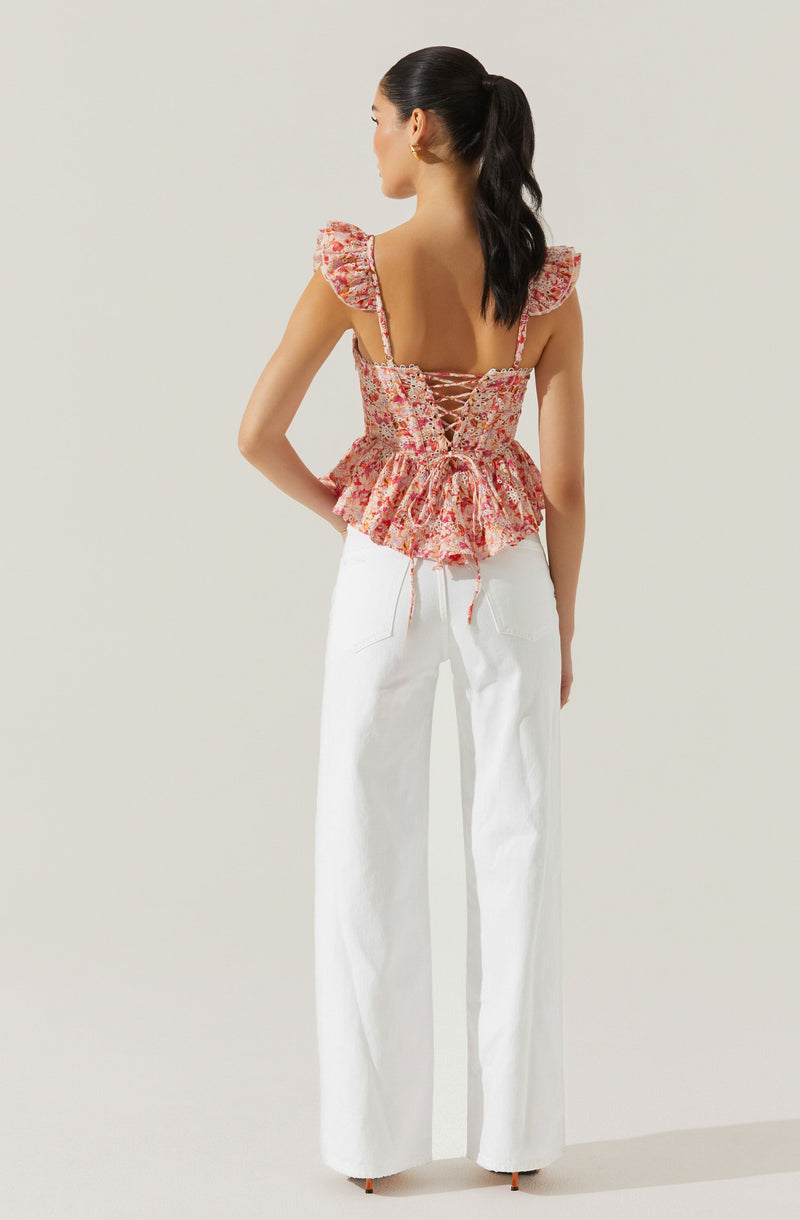 The woman is wearing a sleeveless, floral top with ruffled cap sleeves and a lace-up back design, paired with high-waisted, wide-leg white pants.
