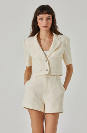 A woman is wearing a cream-colored, textured short-sleeve blazer with a notched lapel collar, two large front pockets, and two shiny buttons. She has paired it with matching high-waisted shorts that also have a textured fabric. The outfit is elegant and coordinated in a monochromatic cream or beige tone.