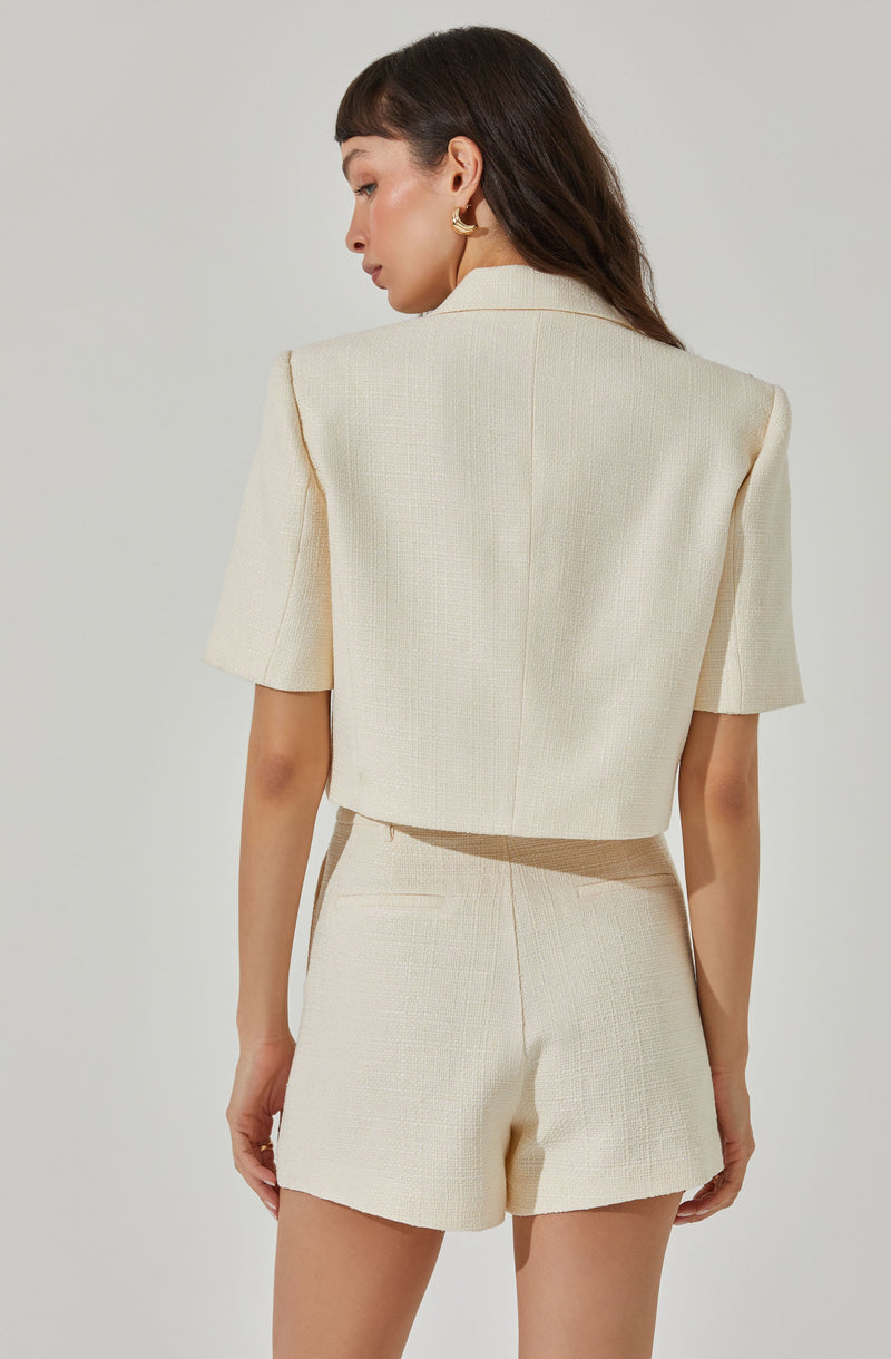 A woman is wearing a cream-colored, textured short-sleeve blazer and matching high-waisted shorts. The blazer has structured shoulders and a classic collar, while the shorts have a tailored fit with a single back pocket. She is accessorizing with gold hoop earrings.
