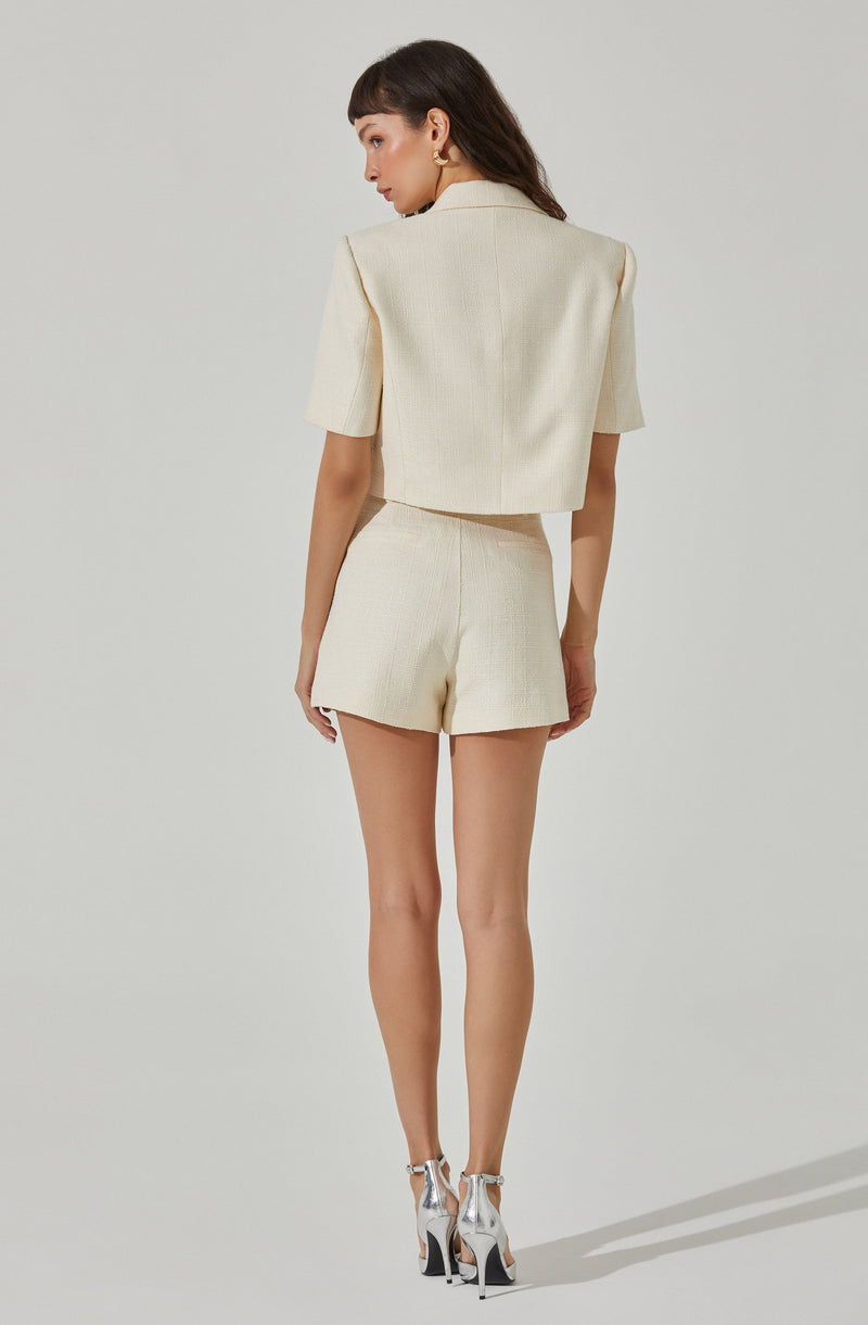 A woman is wearing a cream-colored, textured two-piece outfit consisting of a cropped blazer with short sleeves and a pair of high-waisted shorts. She has on shiny silver high-heeled shoes with ankle straps and is accessorized with gold hoop earrings.