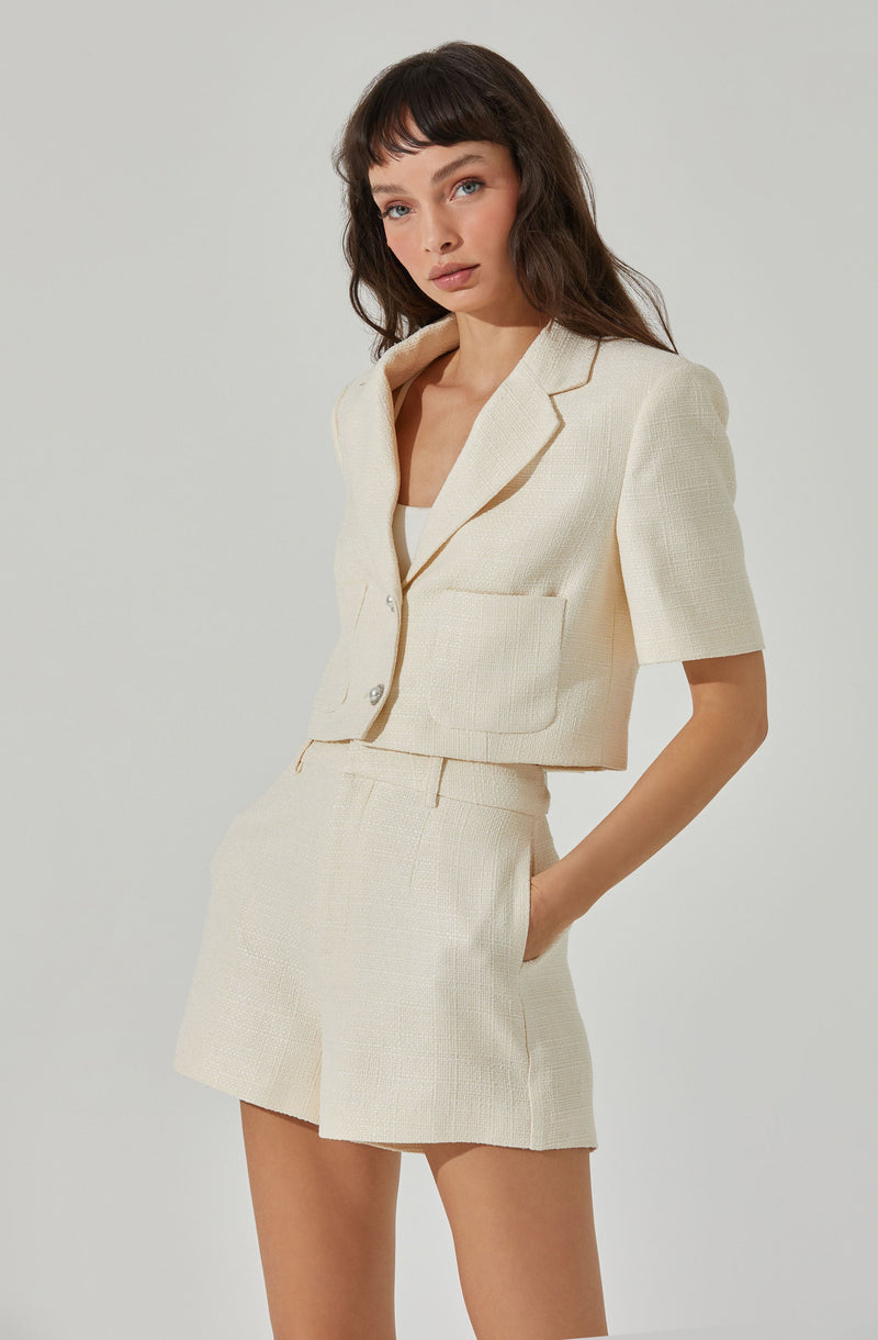 A woman is wearing a cream-colored, textured ensemble consisting of a short-sleeved blazer with a notched lapel, a front pocket, and buttons, paired with matching high-waisted shorts that have a subtle waistband and side pockets.