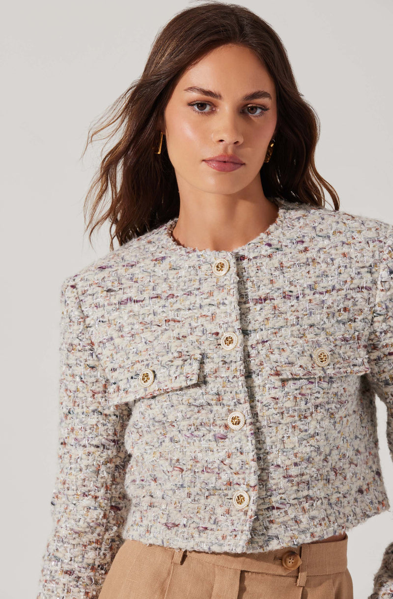 A woman is wearing a textured, multicolored, tweed-style jacket with a round neckline. The jacket features decorative gold buttons down the front and on the flap pockets on each side of the chest. She is also wearing tan high-waisted pants.