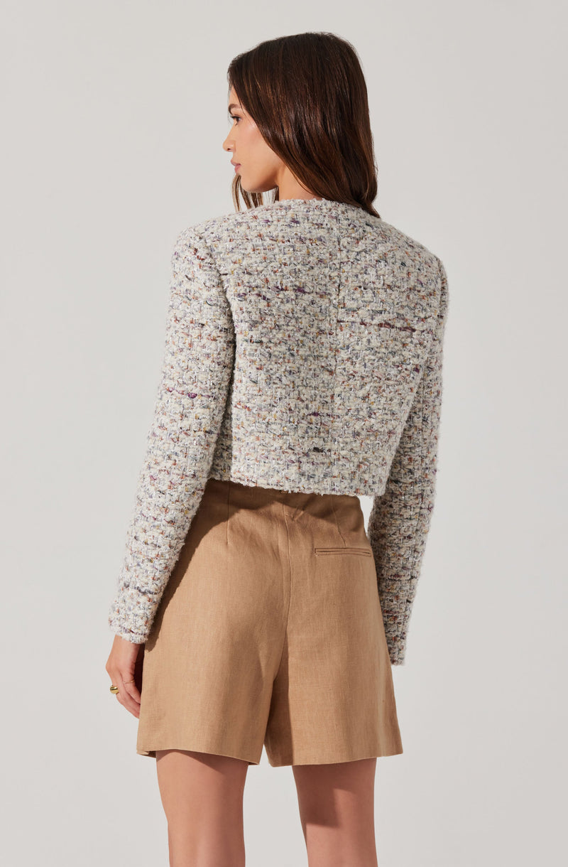 A woman is wearing a textured, multicolored cropped jacket with a boxy fit. The jacket appears to be made of a tweed-like fabric featuring a mix of various colors. She is also wearing high-waisted, light brown shorts with a tailored fit and a back pocket visible.