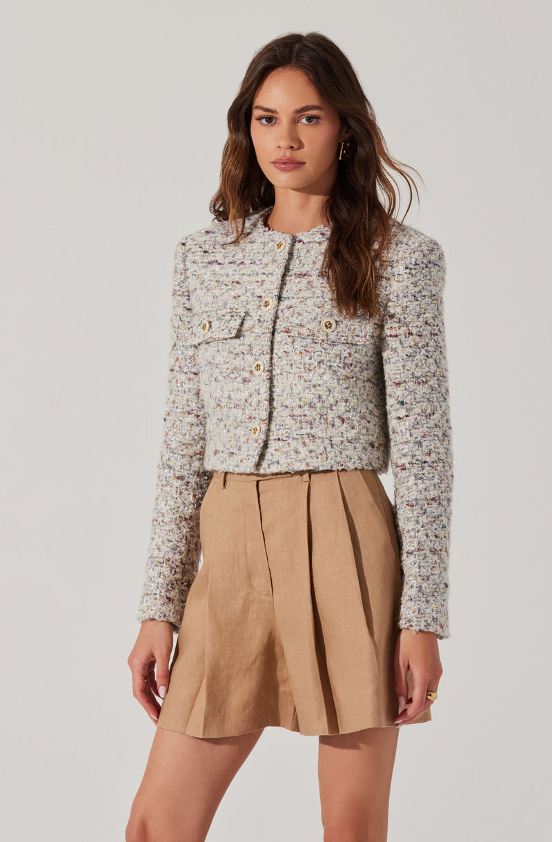 A woman is wearing a textured, multicolored woven cropped jacket with a round neckline and long sleeves. The jacket has decorative gold buttons and a front pocket on the left side. She pairs it with a beige, knee-length pleated skirt made of a lightweight fabric.