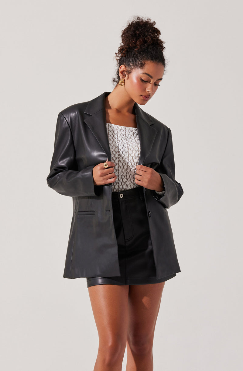 A woman wearing a black leather blazer over a white, textured, short-sleeve top and a black mini skirt. She has curly hair tied up and is accessorized with gold earrings, a gold ring, and a black ring.
