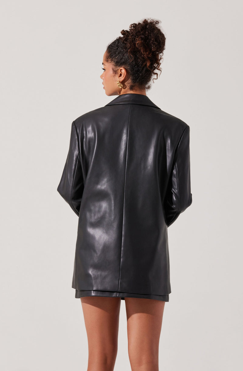 A woman is dressed in a shiny black leather blazer that extends to mid-thigh, with a structured collar and long sleeves. The blazer has a smooth, sleek appearance with a subtle sheen. She is also wearing gold hoop earrings and has her hair styled in a high, voluminous curly ponytail.