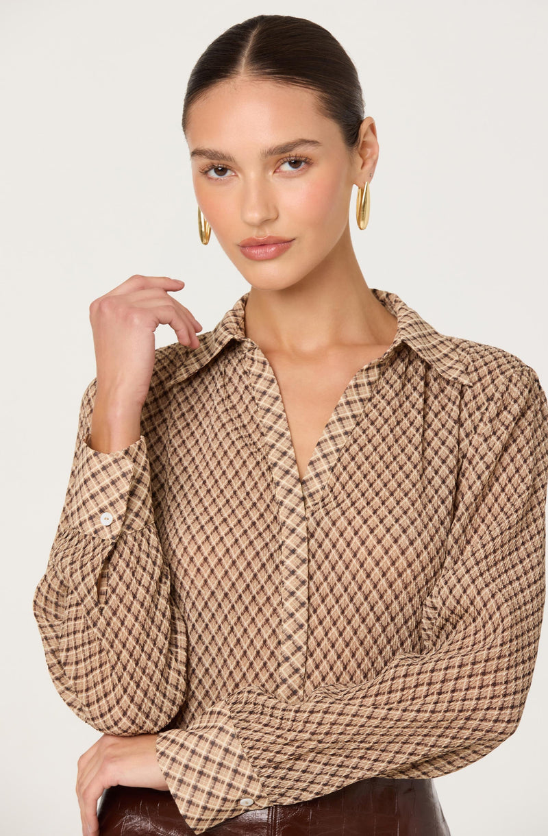 A woman is wearing a long-sleeved button-up shirt with a brown and beige diamond-checkered pattern. She is accessorized with large gold hoop earrings and has her dark hair pulled back in a sleek style.