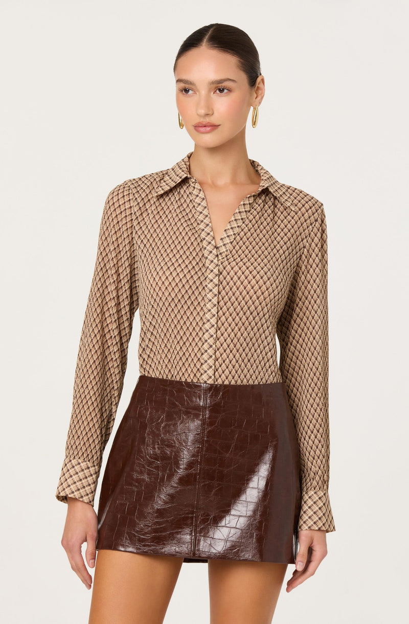 A woman is wearing a long-sleeved button-up shirt with a small diamond pattern in shades of beige, brown, and black. She pairs it with a high-waisted, dark brown leather mini skirt that has a crocodile skin texture.