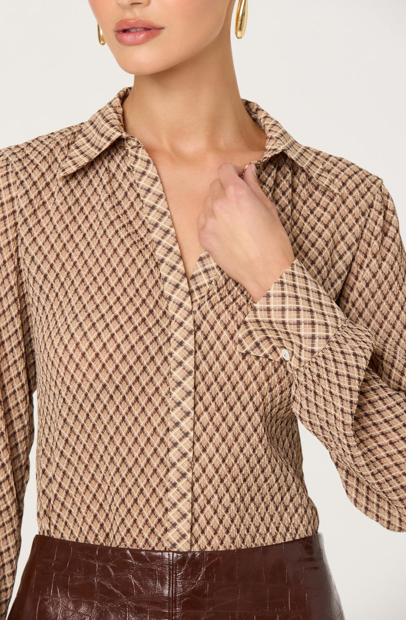 A woman is wearing a long-sleeved button-up shirt with a brown and beige checkered pattern. The shirt has a classic collar and a front pocket on the left side. She has paired it with high-waisted dark brown leather pants.