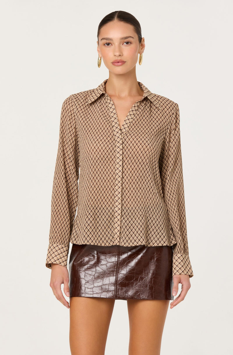A woman is wearing a long-sleeve button-up shirt with a beige and brown geometric diamond pattern, a classic collar, and a relaxed fit. She is also wearing a high-waisted, dark brown, shiny leather mini skirt with a textured, crocodile skin pattern.