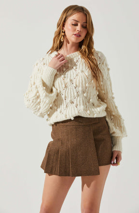 A woman is wearing a cream-colored, chunky knit sweater with a textured, pom-pom detail throughout. She pairs it with a high-waisted, brown plaid pleated skirt. She accessorizes with large, gold drop earrings and rings on her fingers.