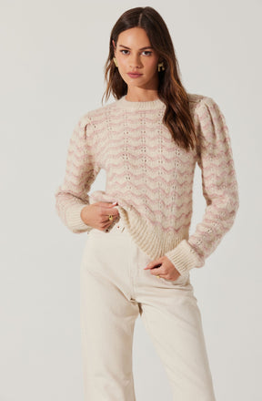 A woman is wearing a cozy, oversized sweater with a soft, light pink and cream zigzag pattern. The sweater has puffed sleeves and a ribbed neckline, cuffs, and hem. She pairs it with high-waisted, off-white pants. The overall look is casual and stylish.