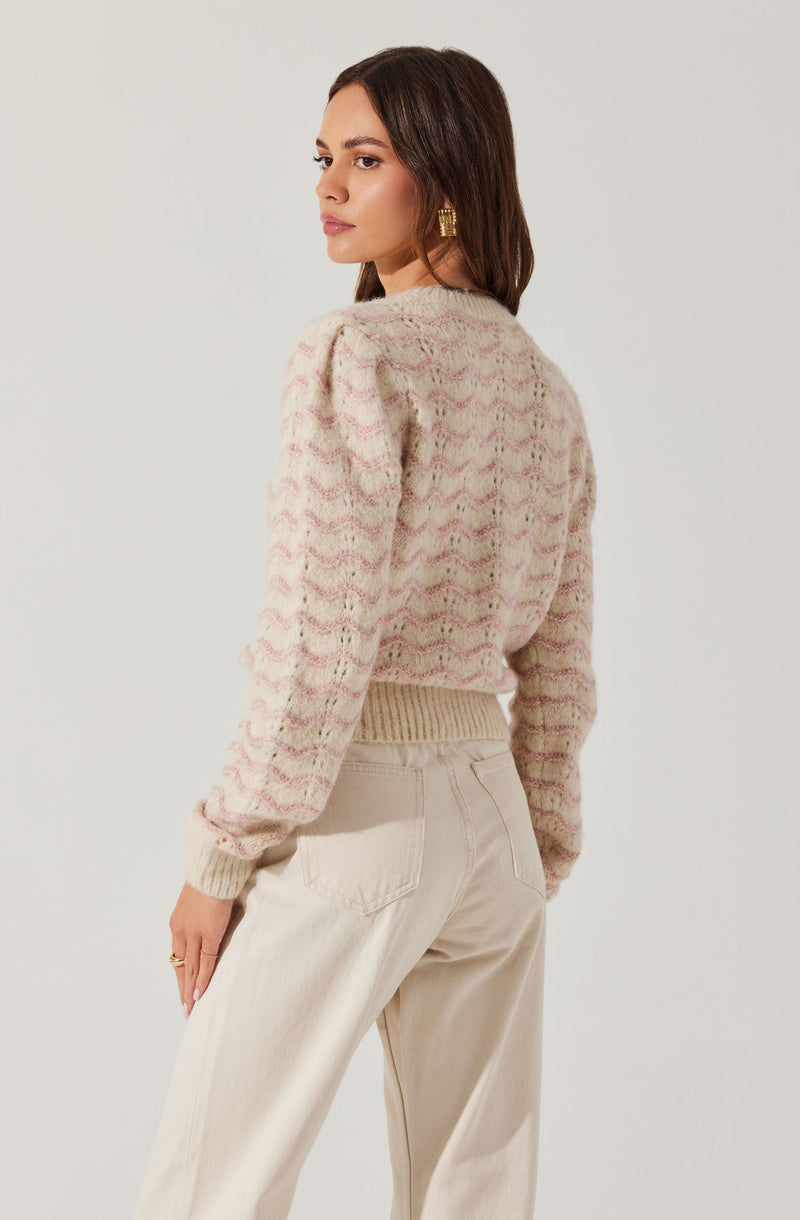 A woman is wearing a textured, long-sleeved sweater with a soft, marbled pattern in shades of pink and cream. The sweater has a ribbed waistband and cuffs, and slightly puffed shoulders. She pairs it with high-waisted, loose-fitting cream-colored pants.