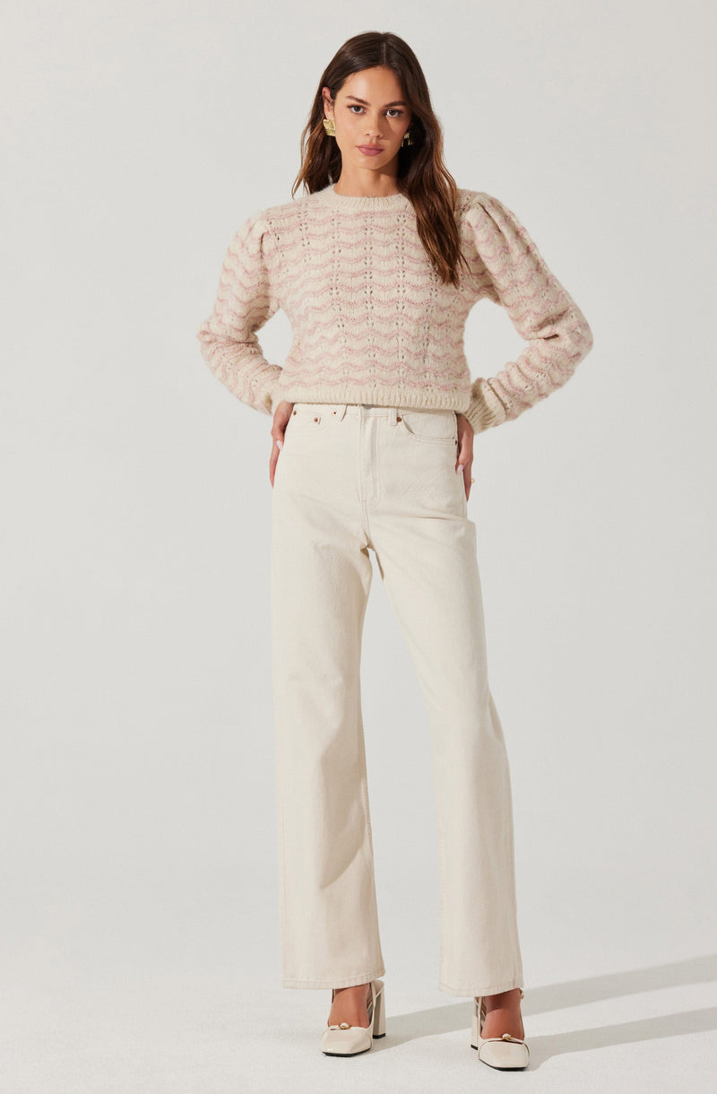 A woman is wearing a pastel pink, chunky knit sweater with puffed long sleeves and a round neckline. She pairs it with high-waisted, off-white wide-leg jeans and white heeled shoes.
