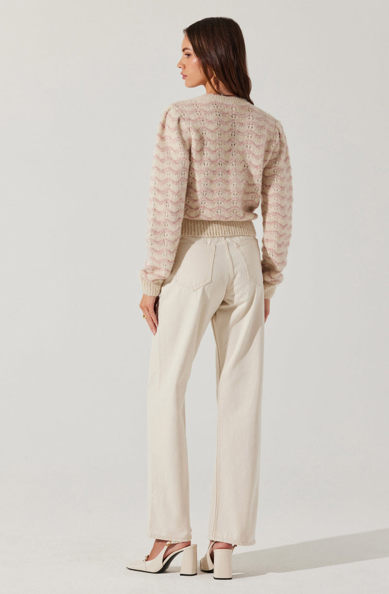 A woman is wearing a soft, multicolored knitted sweater with a pastel pink, beige, and cream pattern and puffed long sleeves. She pairs it with high-waisted, loose-fitting, cream-colored pants and cream high-heeled shoes.