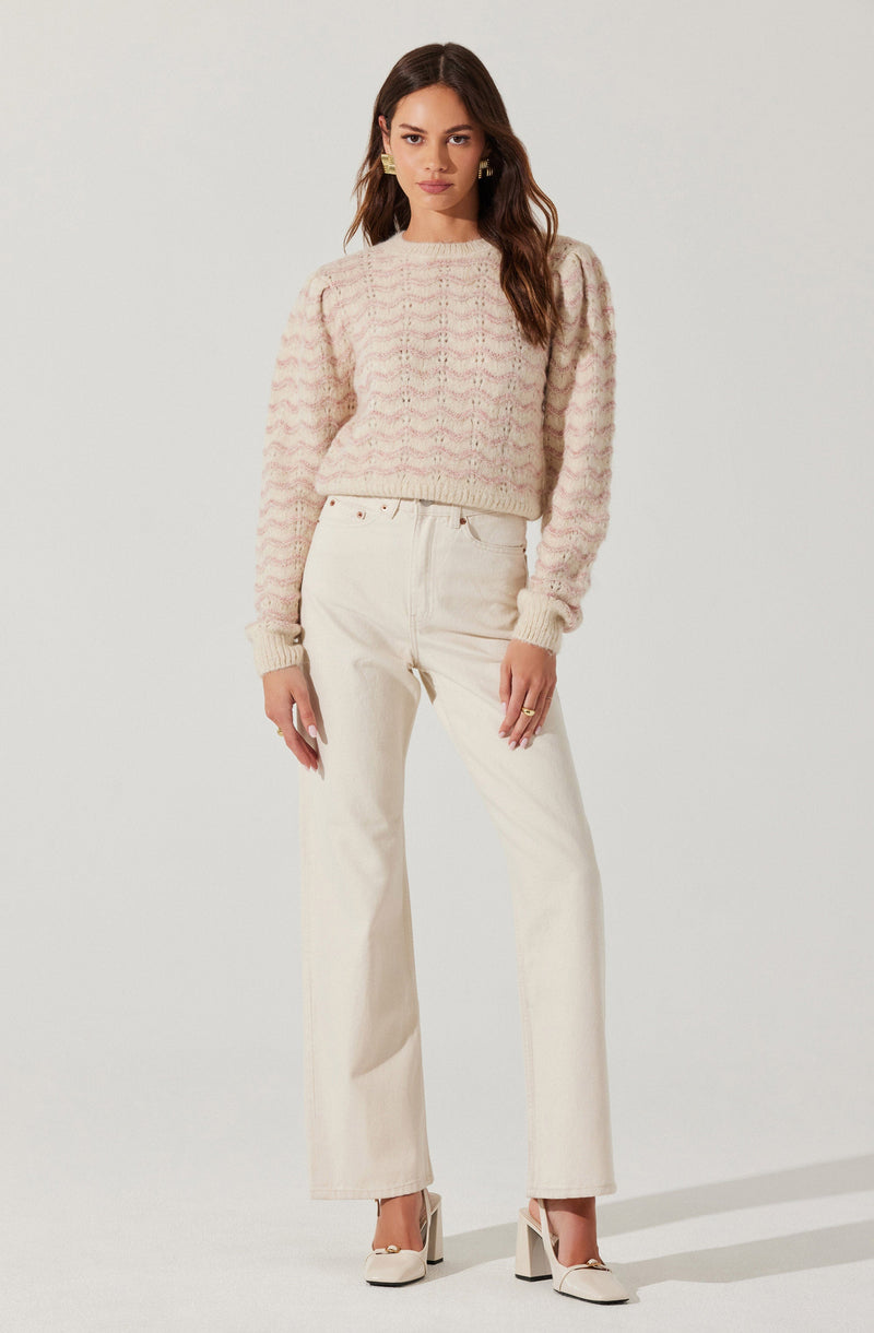 A woman is wearing a light pink and cream-colored knitted sweater with a textured, wave-like pattern. The sweater has long sleeves with ribbed cuffs and a ribbed hem. She is also wearing high-waisted, wide-leg cream-colored pants with a relaxed fit. The outfit is completed with a pair of white high-heeled shoes with an ankle strap. She has long, wavy brown hair, and is accessorized with gold earrings and rings.