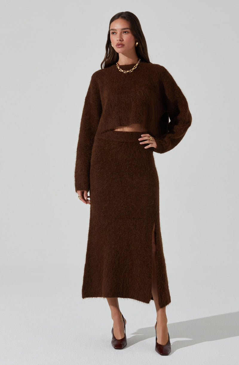 A woman is wearing a long, fuzzy, dark brown knit skirt with a slit on one side, paired with a matching cropped fuzzy sweater that has long sleeves. She accessorized with a chunky gold chain necklace, gold hoop earrings, rings, and dark brown high heels.