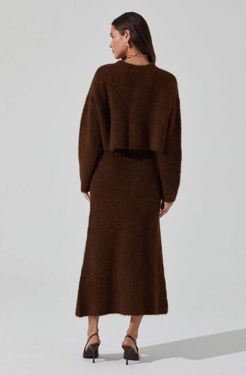 A woman is dressed in a dark brown, fuzzy, long-sleeved top that is cropped at the waist, paired with a matching high-waisted, ankle-length skirt. She is wearing a chunky gold chain necklace and gold hoop earrings. Her footwear consists of dark brown high-heeled sandals with ankle straps.