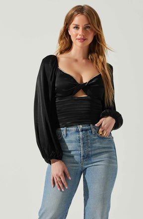 A woman with long, wavy, reddish-brown hair is wearing a black satin long-sleeve top with puffed shoulders, a twisted front detail, and a small cutout below the bust, accented with a rhinestone trim along the neckline. She is also wearing light blue high-waisted jeans with a relaxed fit.