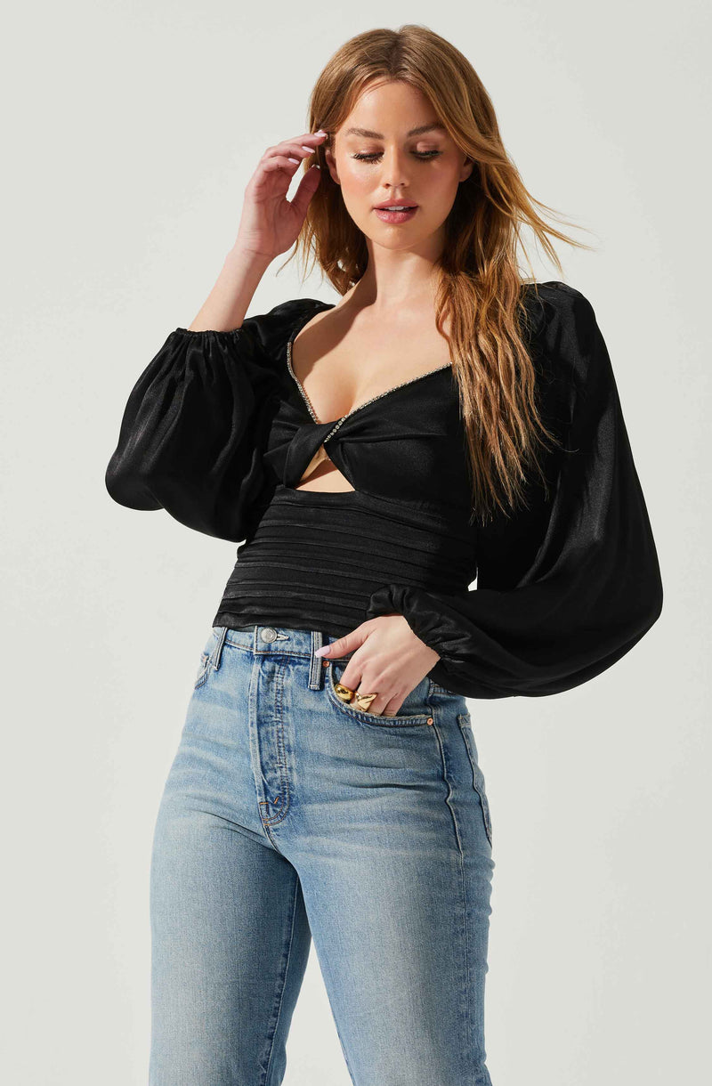 A woman with long, wavy red hair wearing a black satin top with a front zipper, long puffed sleeves, and a fitted waist. She is also wearing high-waisted, light blue jeans.