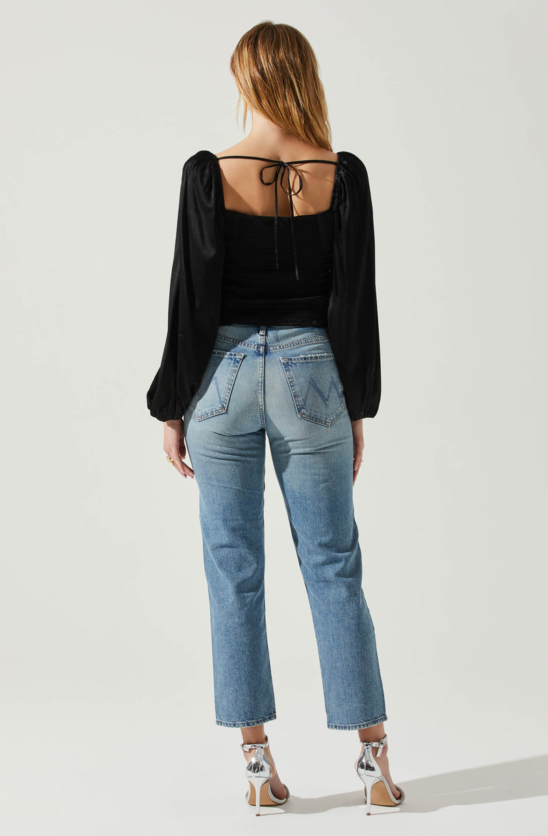 A woman is wearing a black top with long, billowy sleeves, and the top has a square neckline with thin straps tied at the back. She is also wearing high-waisted, light blue jeans with a slightly faded wash and visible stitching on the back pockets. She has on silver high-heeled sandals with ankle straps.