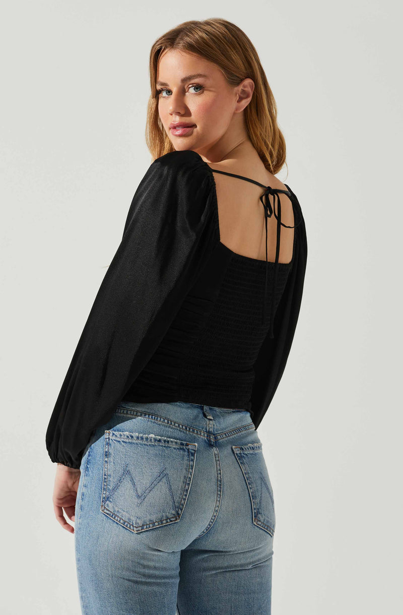 A woman is wearing a black top with a smocked bodice and thin shoulder straps tied at the back. She has on a black, shiny, long-sleeved jacket with elasticized cuffs. She is also wearing light blue high-waisted jeans with back pockets, one of which has a stitched "W" design.