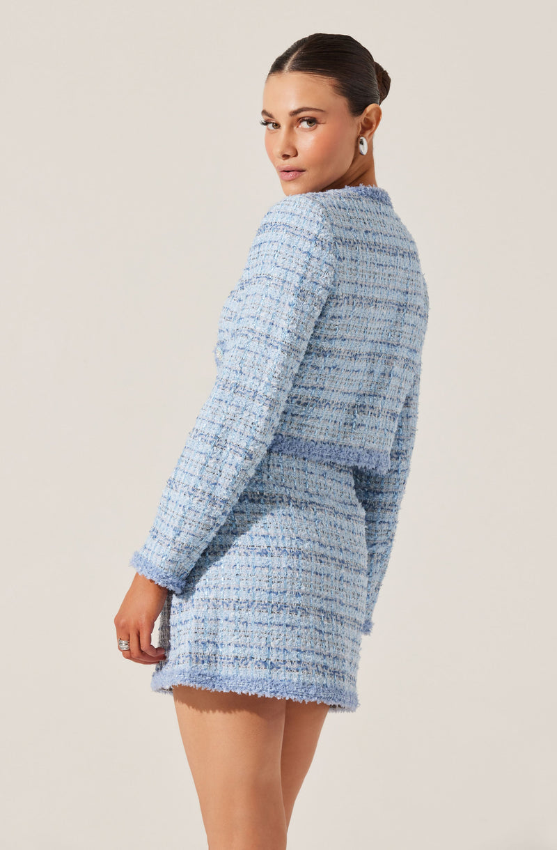 A woman is wearing a light blue, textured, tweed-style blazer and matching skirt set. The blazer has long sleeves and a collarless design, and the skirt is short and fitted. The fabric appears thick and woven, with a mix of light and dark blue threads. The woman has her hair styled in a sleek bun and is wearing earrings.