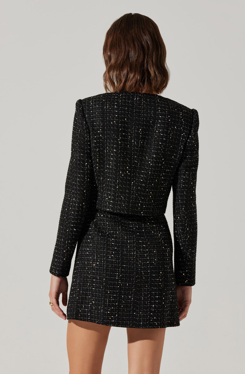A woman is wearing a matching black tweed blazer and skirt set. The blazer has structured shoulders and a textured pattern with tiny gold accents throughout. The skirt is a high-waisted, straight style that matches the blazer's fabric and pattern. The outfit is tailored and elegant, with a classic, sophisticated look.