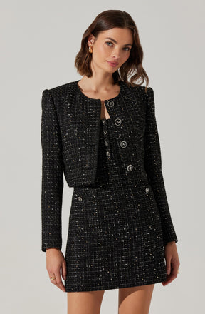 A woman is wearing a black, textured, tweed-style jacket and matching high-waisted skirt. The jacket is cropped with a rounded neckline, and both the jacket and skirt feature a subtle checkered pattern with small metallic threads. The jacket has long sleeves and is fastened with large black buttons down the front.