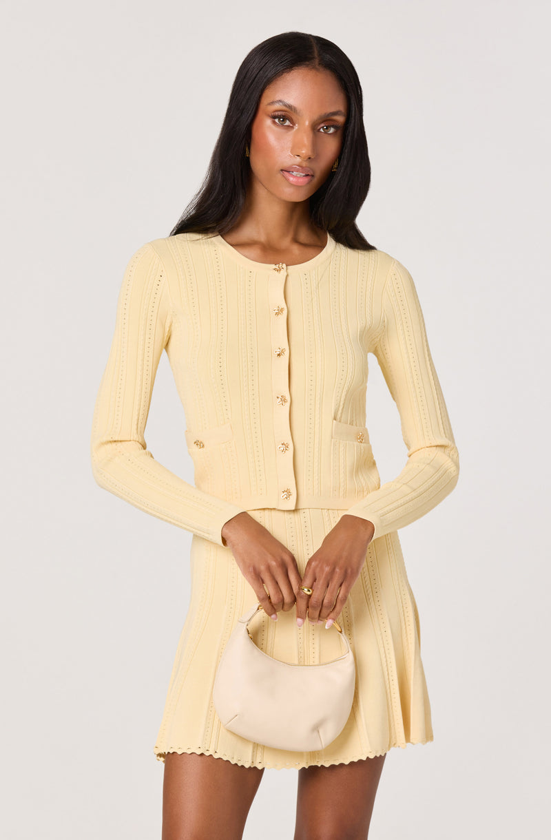Woman wearing a pale yellow knit cardigan and matching skirt, holding a cream handbag.