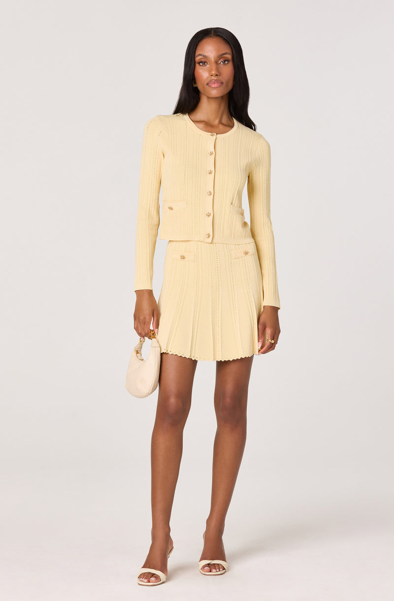 Woman wearing a pale yellow, knit long-sleeve cardigan with buttons and matching short skirt.
