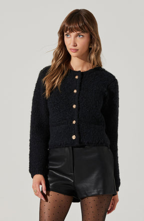A woman is wearing a black, textured, long-sleeve button-up cardigan with gold buttons, paired with high-waisted black leather shorts. She has on black sheer tights with small polka dots.