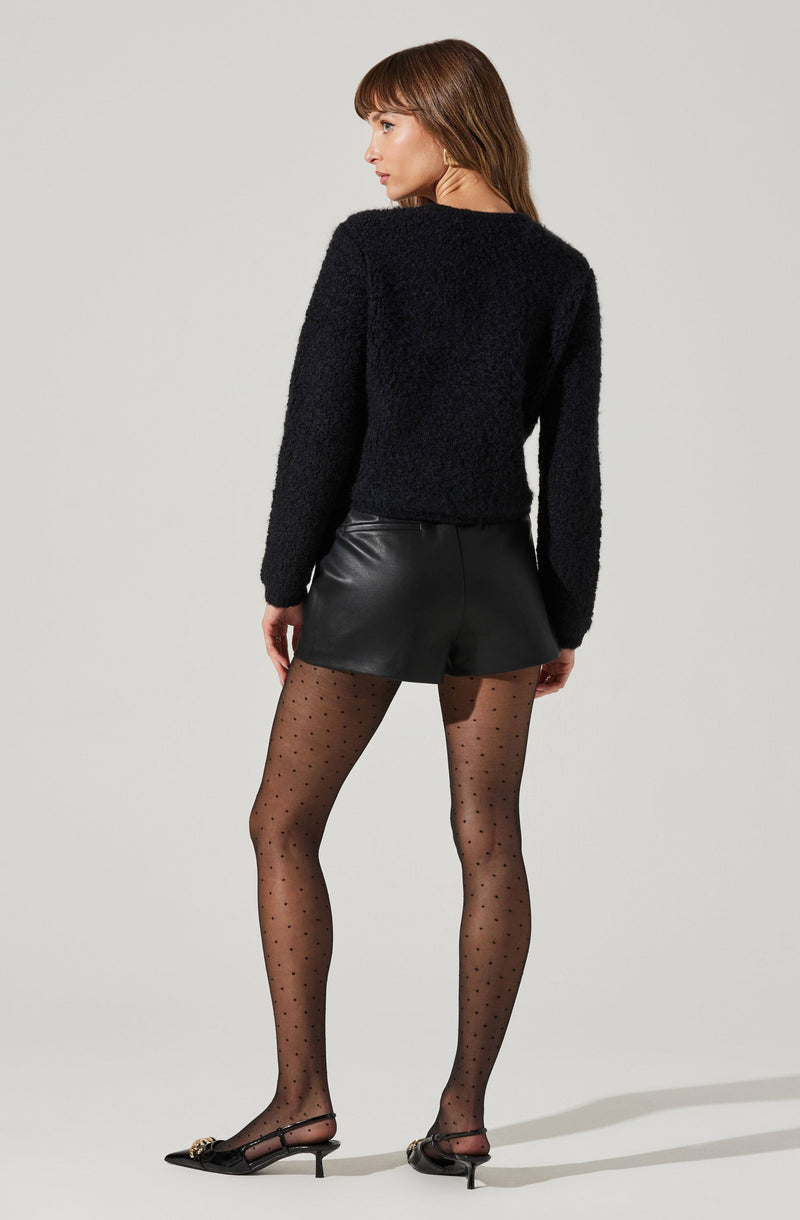 A woman is wearing a black, fuzzy long-sleeve sweater, black leather shorts, sheer black tights with small polka dots, and black high-heeled shoes with ankle straps and decorative bow details.