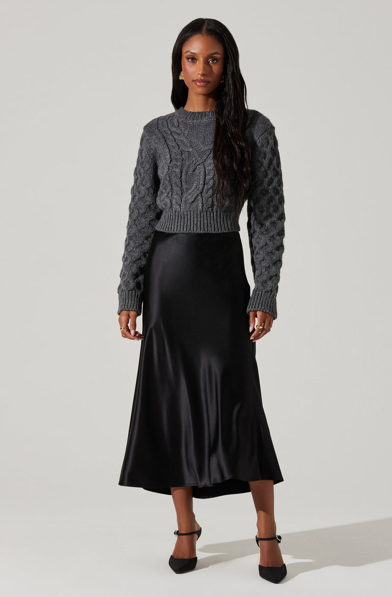 A woman is wearing a dark gray chunky knit sweater with cable knit patterns on the front and sleeves, paired with a long, black satin or silk midi skirt that has a smooth, shiny finish. She is also wearing black pointed-toe heels with ankle straps.