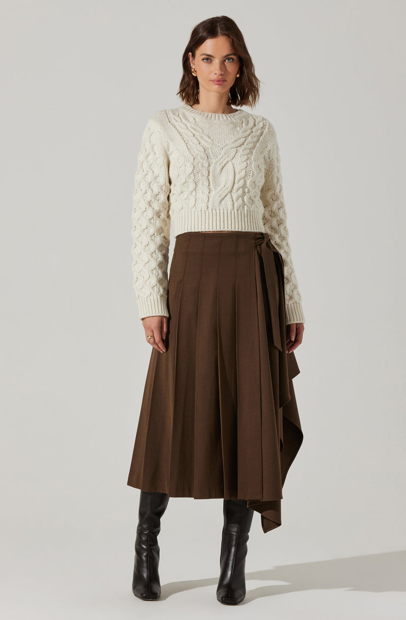 A woman is wearing a cream-colored, cable-knit sweater with a round neckline and long sleeves. She pairs it with a high-waisted, brown pleated skirt that features a wrap detail with a tie on the side. She completes her outfit with black knee-high boots.