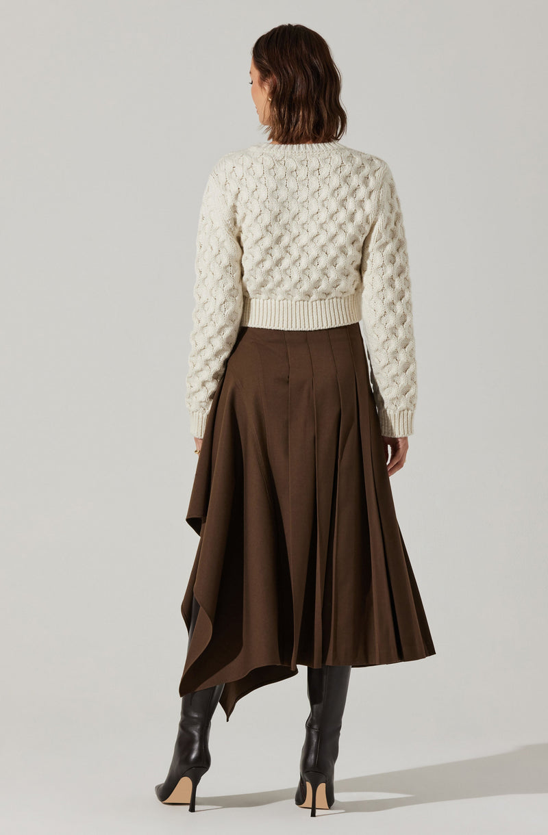 A woman is wearing a cream-colored knit sweater with a textured, cable-knit pattern, a wide ribbed hem, and long sleeves. She pairs it with a high-waisted, brown pleated skirt that falls below the knees, along with black high-heeled boots.