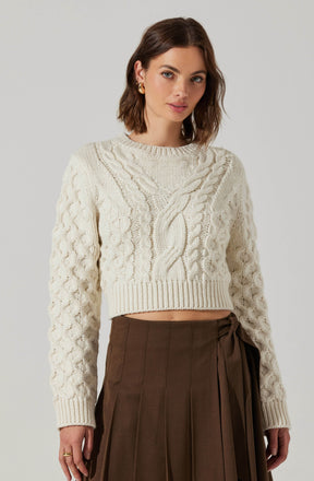 A woman is wearing a cream-colored, cropped cable knit sweater with a round neckline and intricate cable patterns on the front and sleeves. She pairs it with a high-waisted brown pleated skirt that features a side wrap detail with a tied bow.
