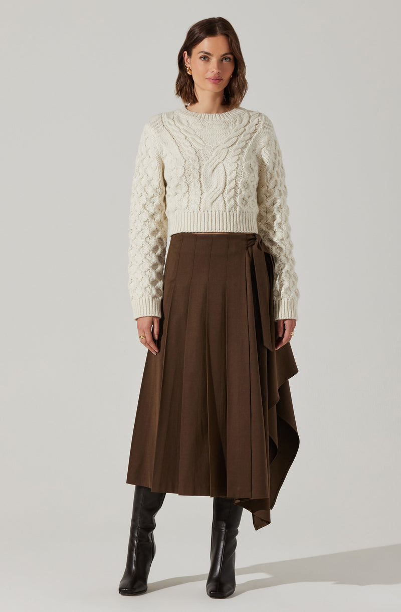A woman is wearing a cream-colored, chunky cable-knit sweater with a round neckline and long sleeves. She pairs it with a high-waisted, brown pleated midi skirt that features an attached, asymmetrical draped panel on one side. She completes her outfit with black knee-high boots.