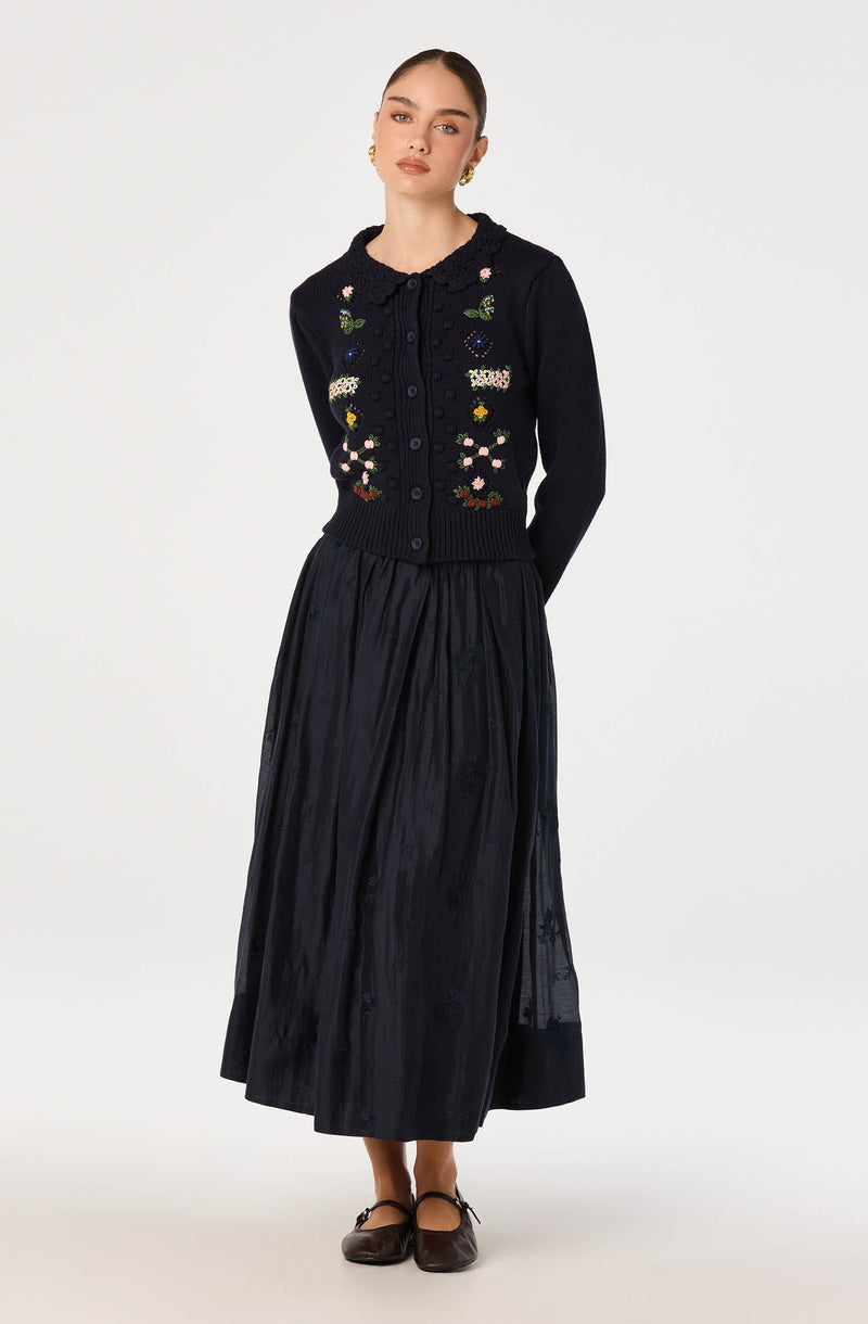 Woman wearing a navy blue embroidered cardigan with floral patterns and a matching long, pleated navy skirt.