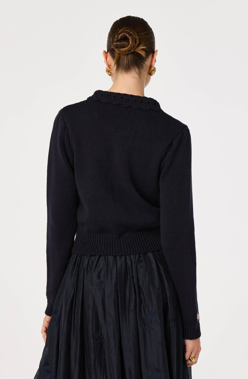 Woman wearing a black knitted sweater with a scalloped collar and a shiny black skirt with embroidery.