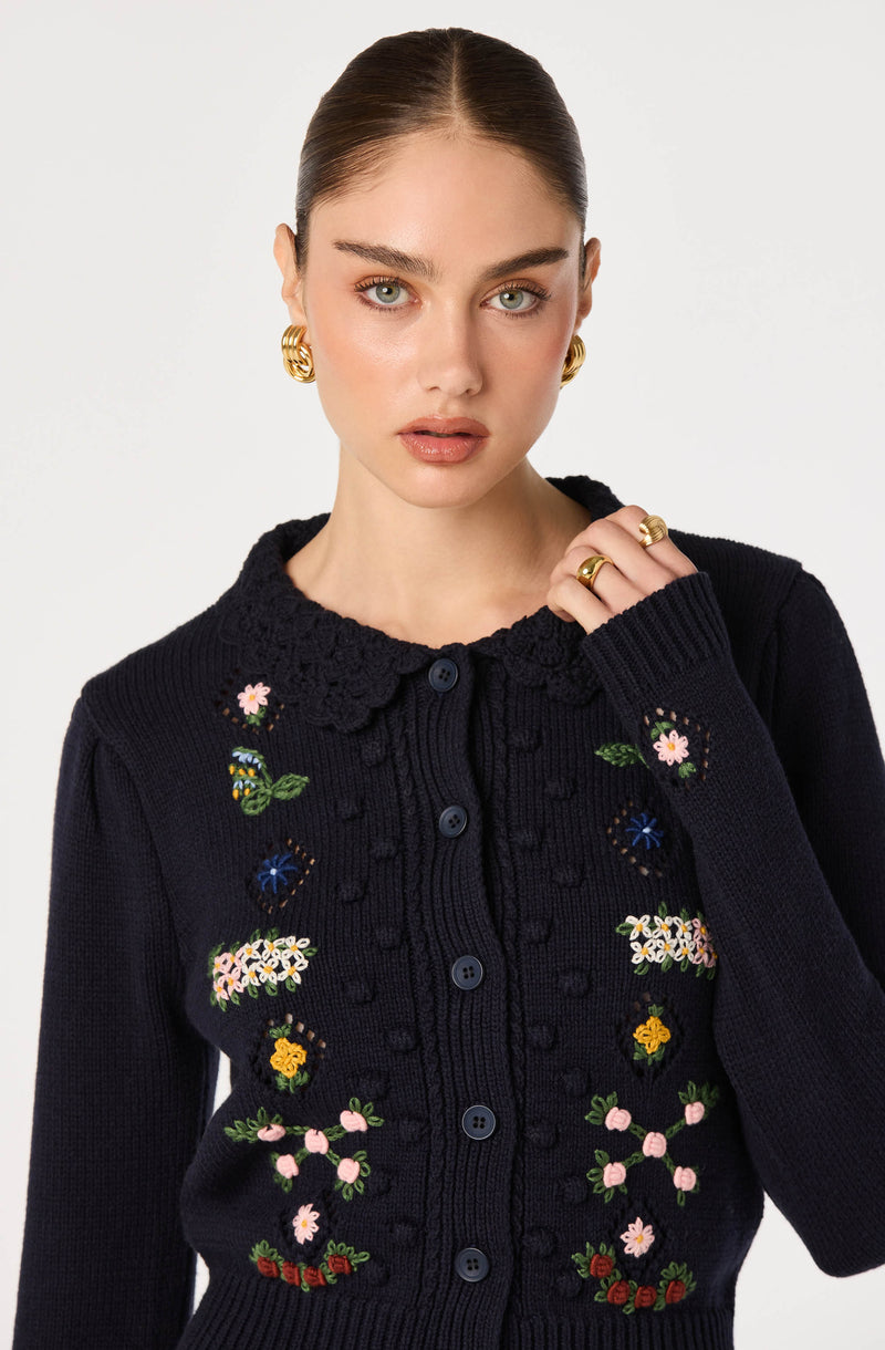 Woman wearing a navy blue knit cardigan with colorful floral embroidery and a scalloped collar.