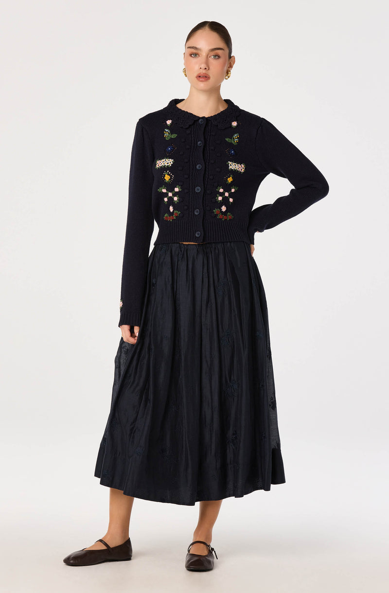 Woman wearing a black cardigan with colorful floral embroidery and a long black skirt.