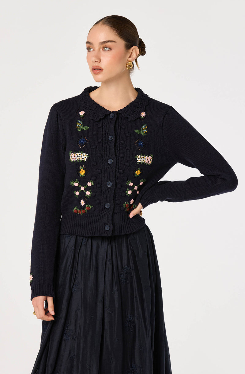 Woman wearing a black knitted cardigan with floral embroidery and a black satin skirt.