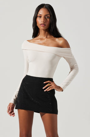 A woman is wearing an off-the-shoulder, long-sleeved white top with a wide neckline that drapes over her shoulders. She pairs it with a high-waisted, black mini skirt that has a shiny, textured surface.